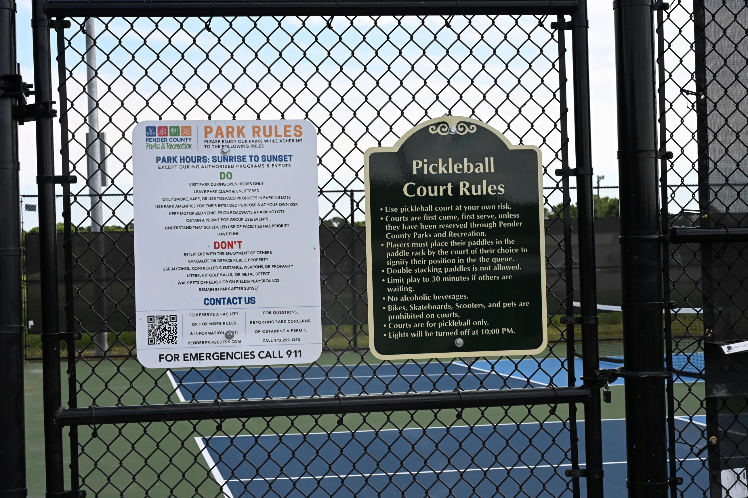 Signs on a chain-link fence detail park and pickleball court rules. The background shows a pickleball court with blue and green surfaces.