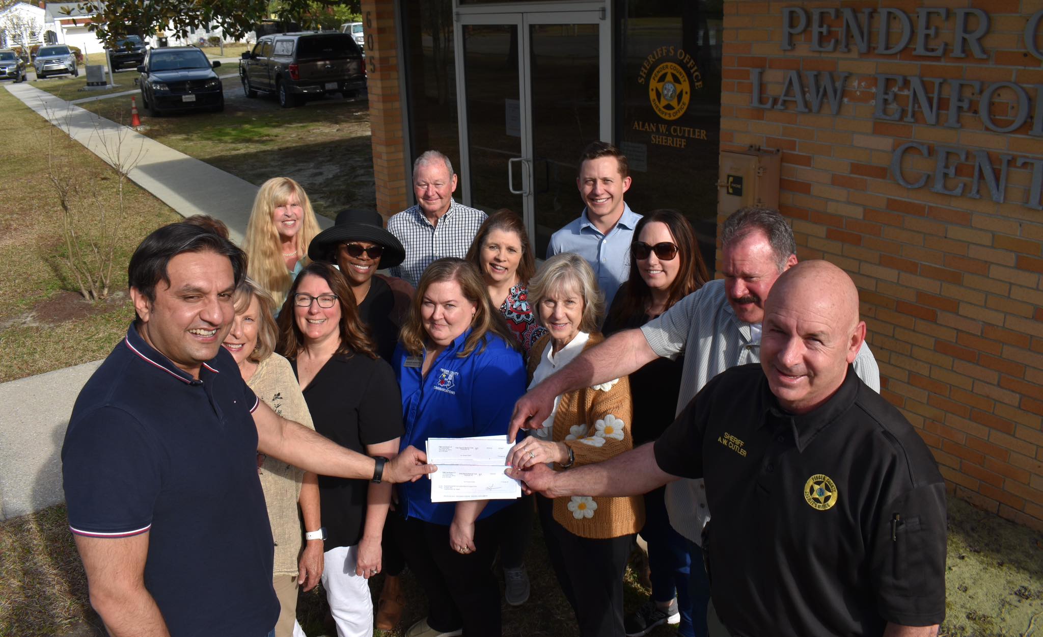 A group of smiling people stand outside the Pender County Law Enforcement Center. Two individuals in front hold a large check