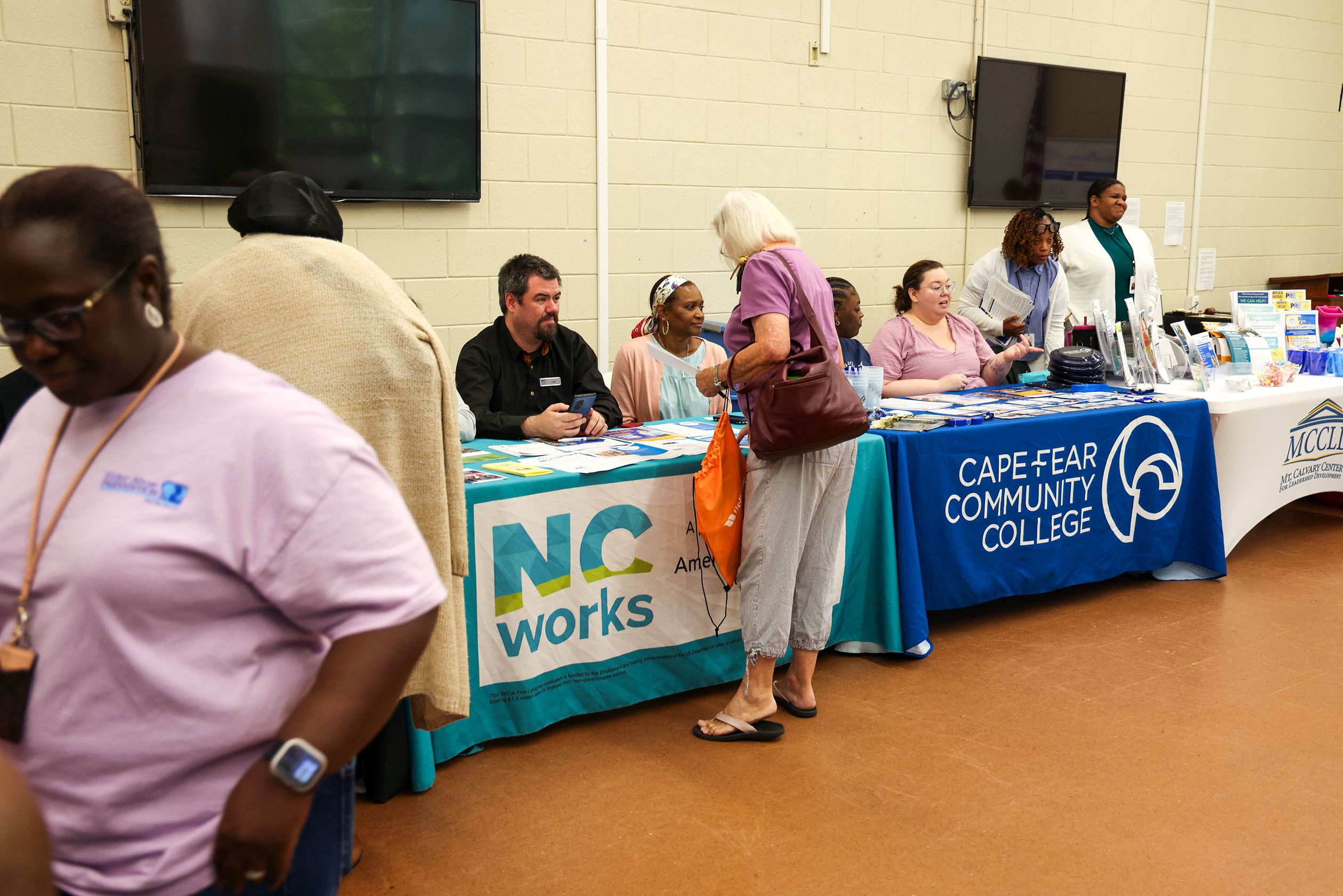 People interact at a vendor fair, with tables for NCWorks and Cape Fear Community College. Information and resource materials are displayed.